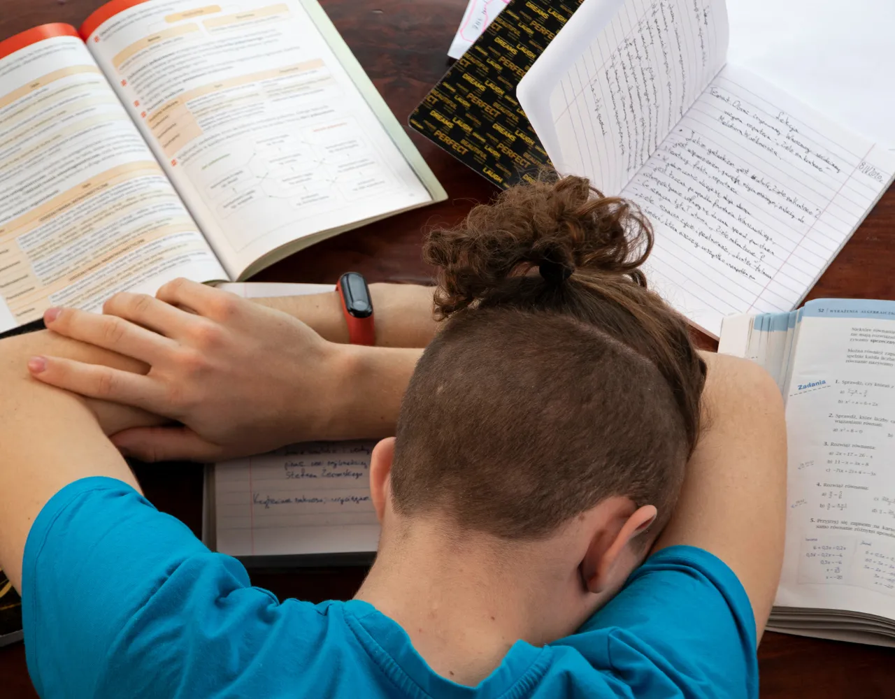 Pessoa de cabelo curto e camisa azul apoiando a cabeça sobre os braços cruzados sobre um caderno aberto, rodeado de livros e papéis abertos, parecendo estar estudando.