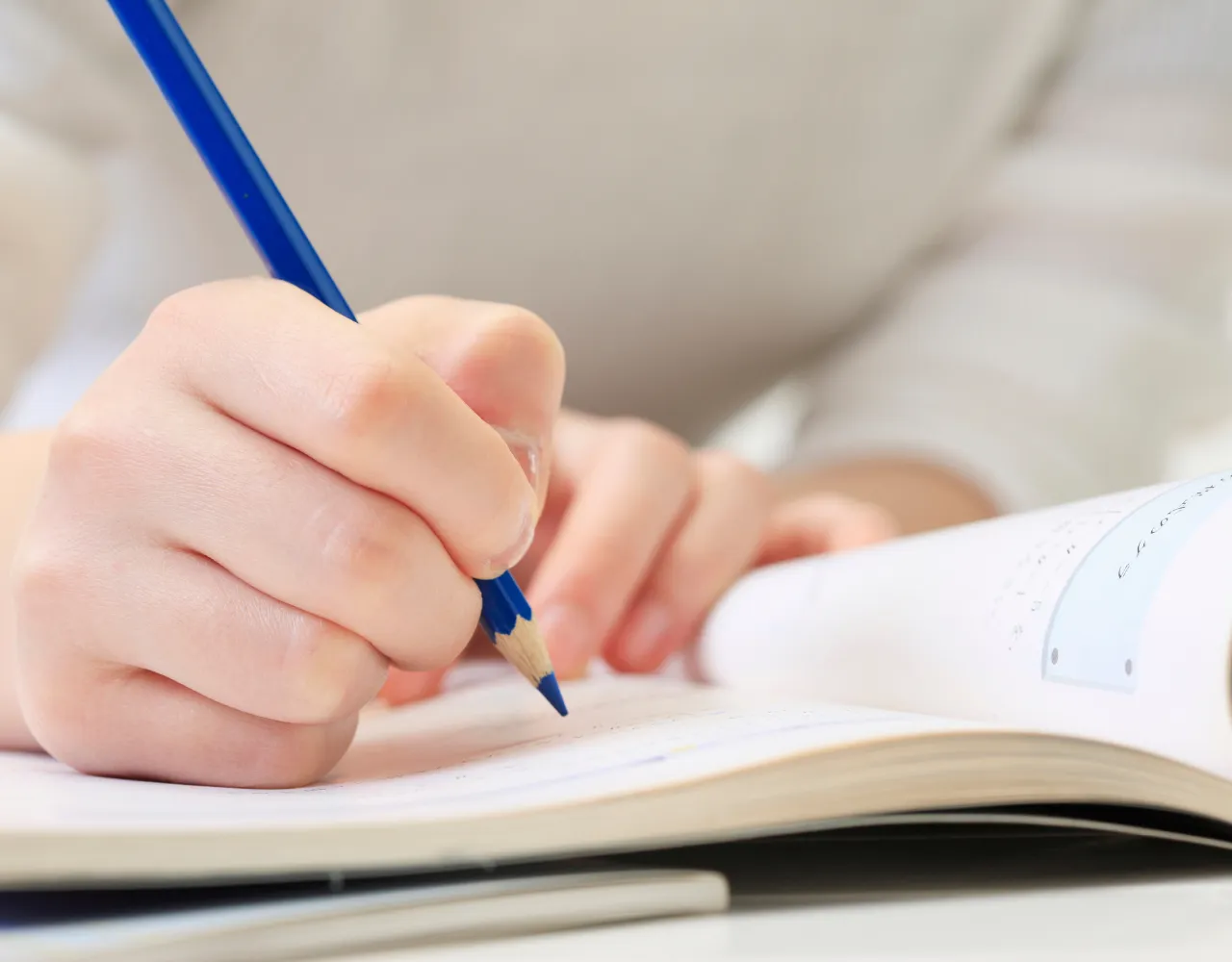 A mão de uma pessoa segurando uma caneta azul está escrevendo em um caderno aberto sobre uma mesa.