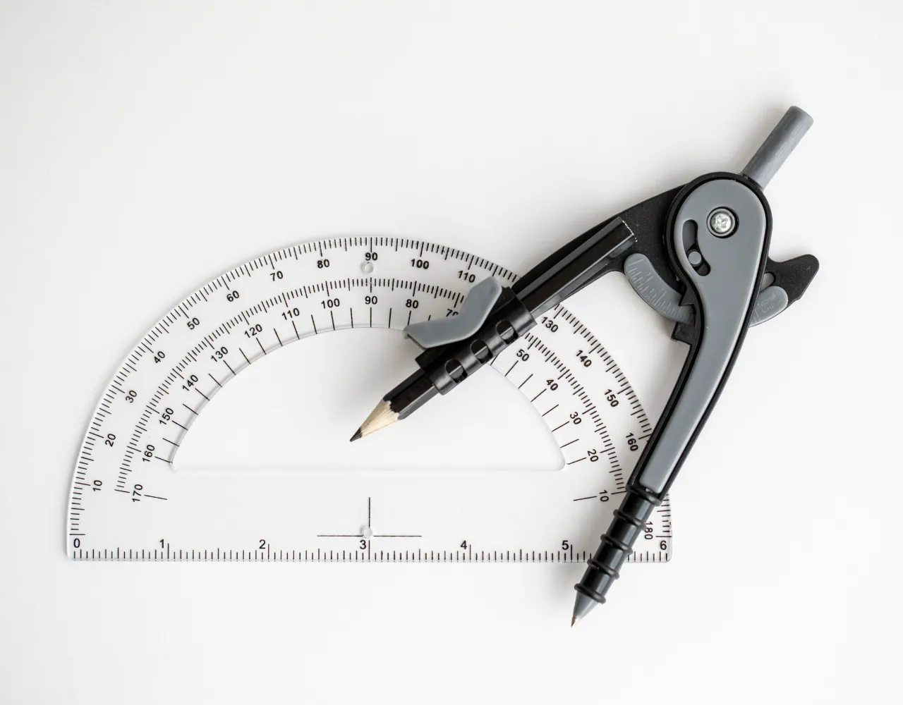 Um transferidor de plástico transparente marcado com graus e radianos é colocado ao lado de uma bússola de metal preto com um lápis preso, sobre fundo branco.