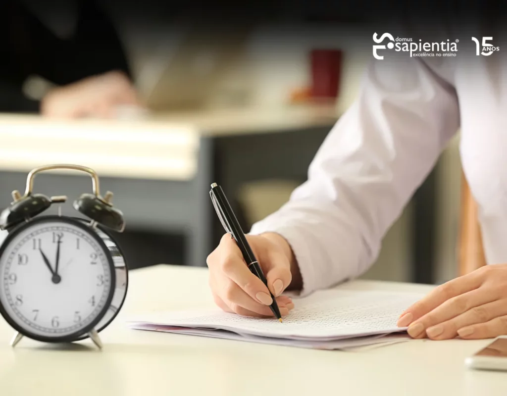 Pessoa de camisa branca escrevendo em papel em uma mesa com um despertador em primeiro plano. Logotipo "Sapientia" visível no canto superior direito.