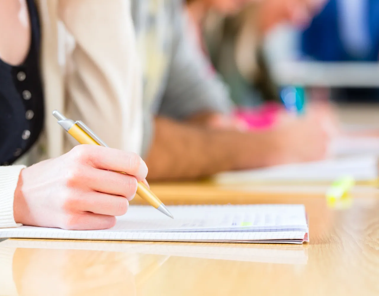 Close de uma pessoa escrevendo em um caderno com uma caneta sobre uma mesa, com outras pessoas no fundo desfocado focadas em seu trabalho.