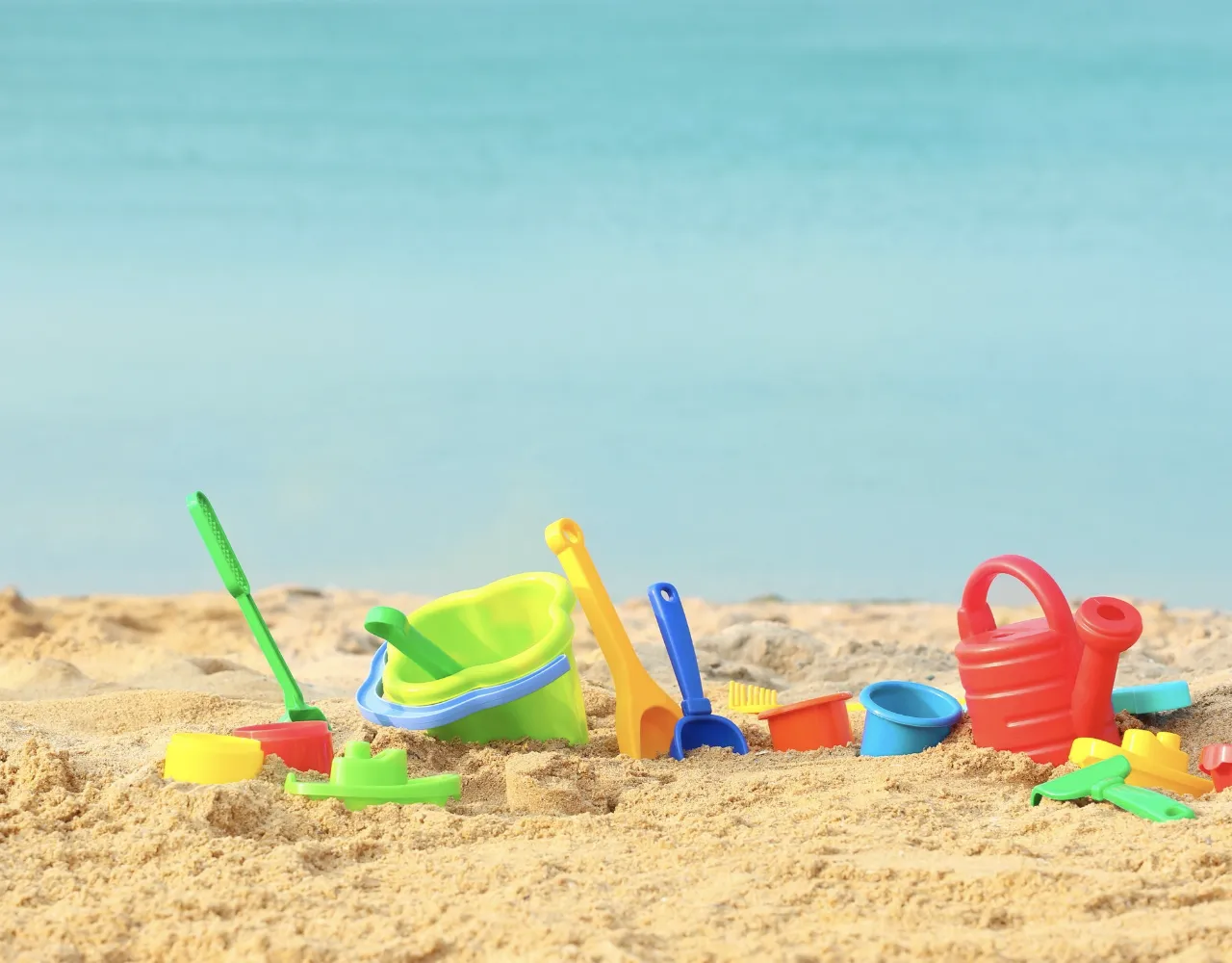 Brinquedos de praia coloridos, incluindo baldes, pás e um regador, estão espalhados na praia com mar azul calmo ao fundo.