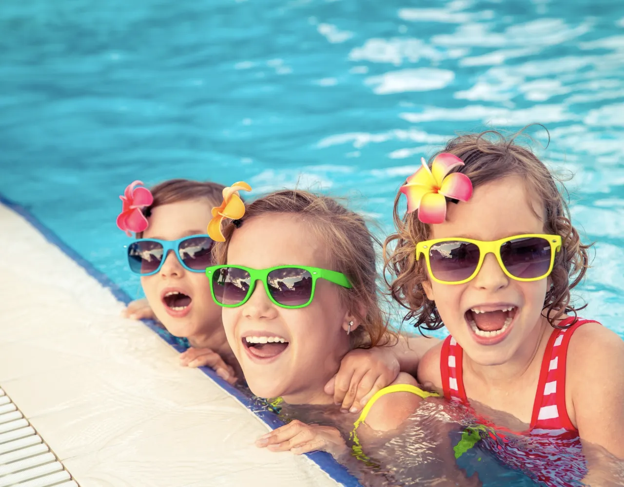 Três crianças usando óculos escuros coloridos e presilhas de cabelo com flores estão apoiadas na beira de uma piscina, sorrindo e rindo.