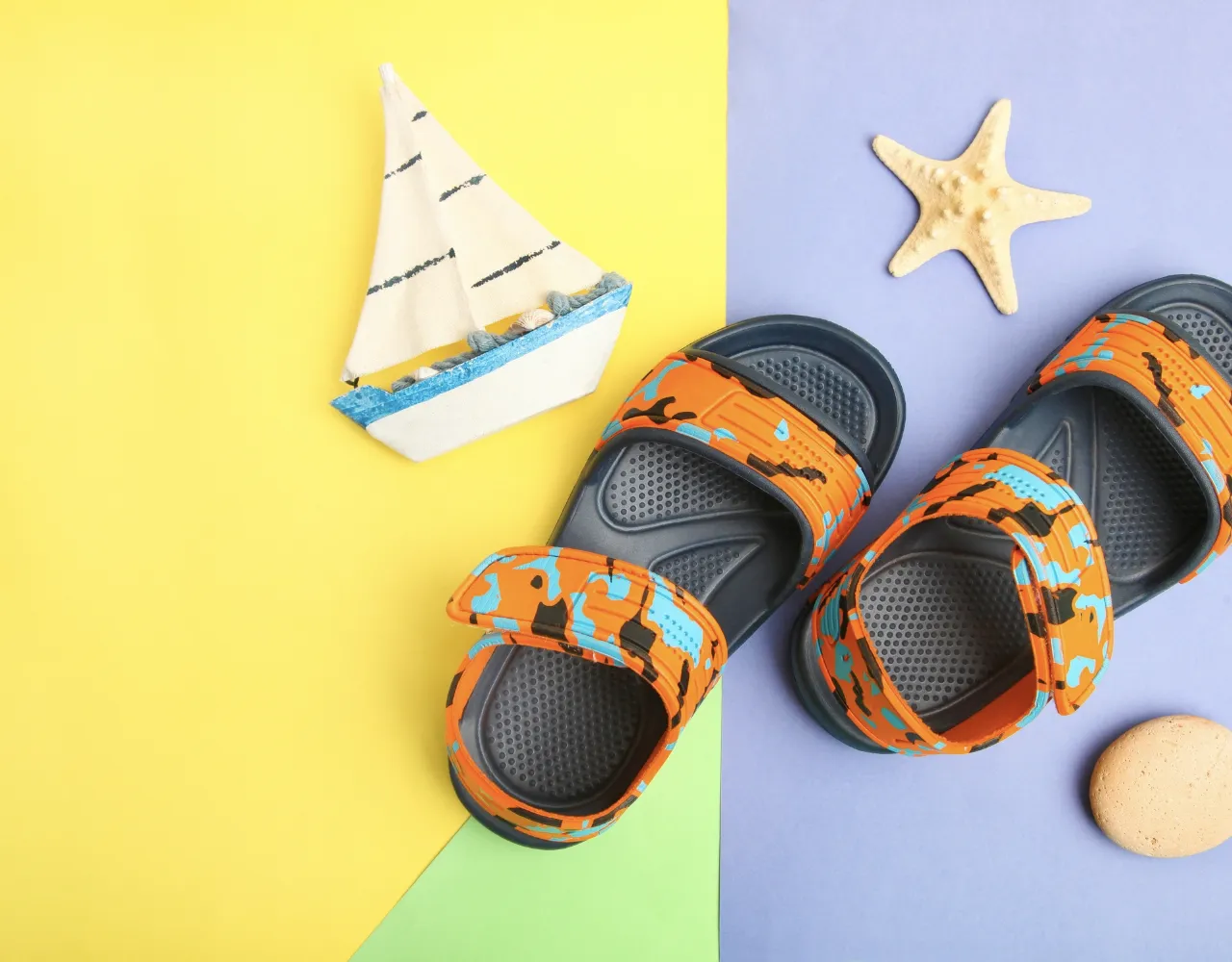 Um par de sandálias infantis laranja e azuis sobre um fundo colorido com um veleiro de brinquedo e uma estrela do mar.