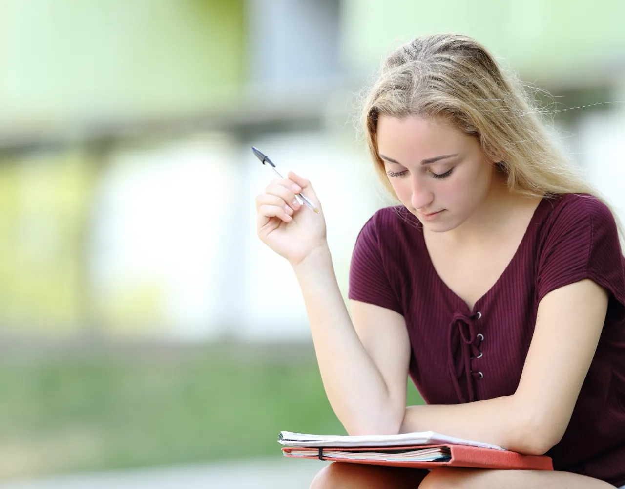 Uma jovem com longos cabelos loiros segura uma caneta e olha para um caderno aberto enquanto está sentada ao ar livre em um dia ensolarado.
