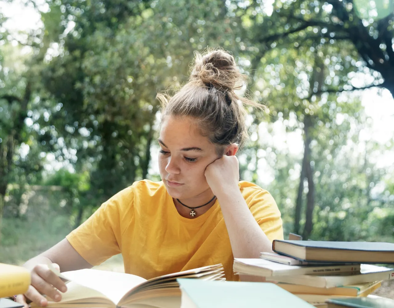 Uma pessoa com penteado em coque e camisa amarela lê um livro em uma mesa ao ar livre cercada por mais livros, com árvores ao fundo.