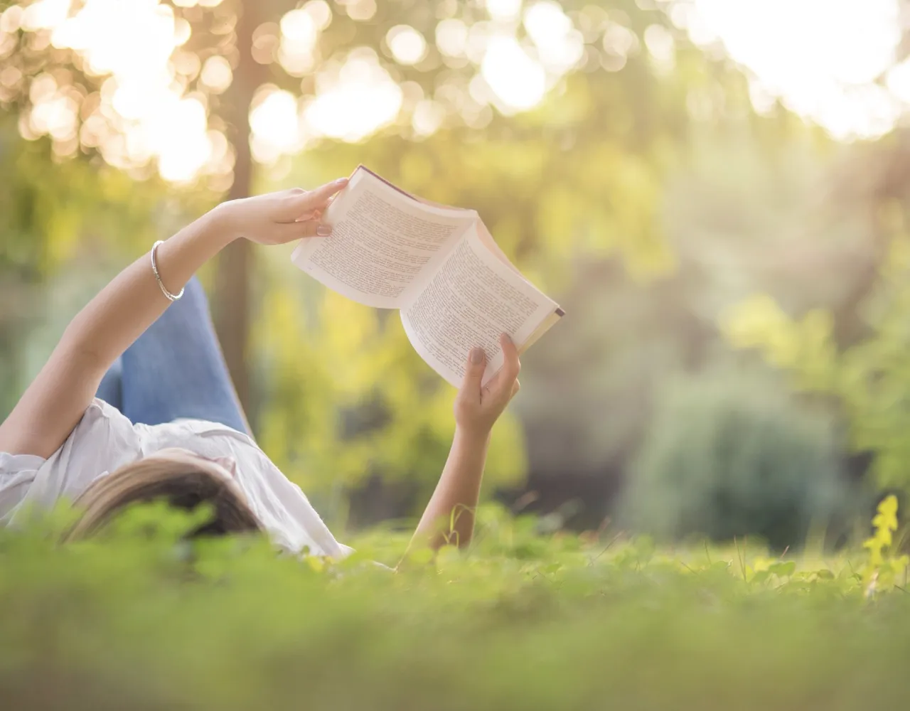 Pessoa deitada na grama de um parque, segurando e lendo um livro aberto com a luz do sol filtrada pelas árvores ao fundo.