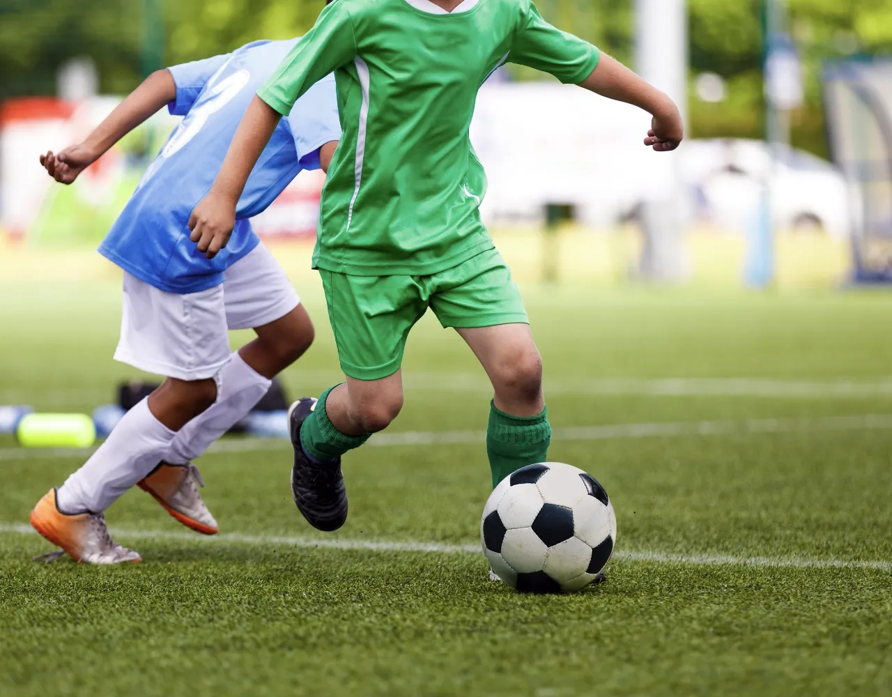 Duas crianças jogando futebol, uma de uniforme verde driblando a bola e outra de uniforme azul e branco correndo logo atrás em um campo gramado.