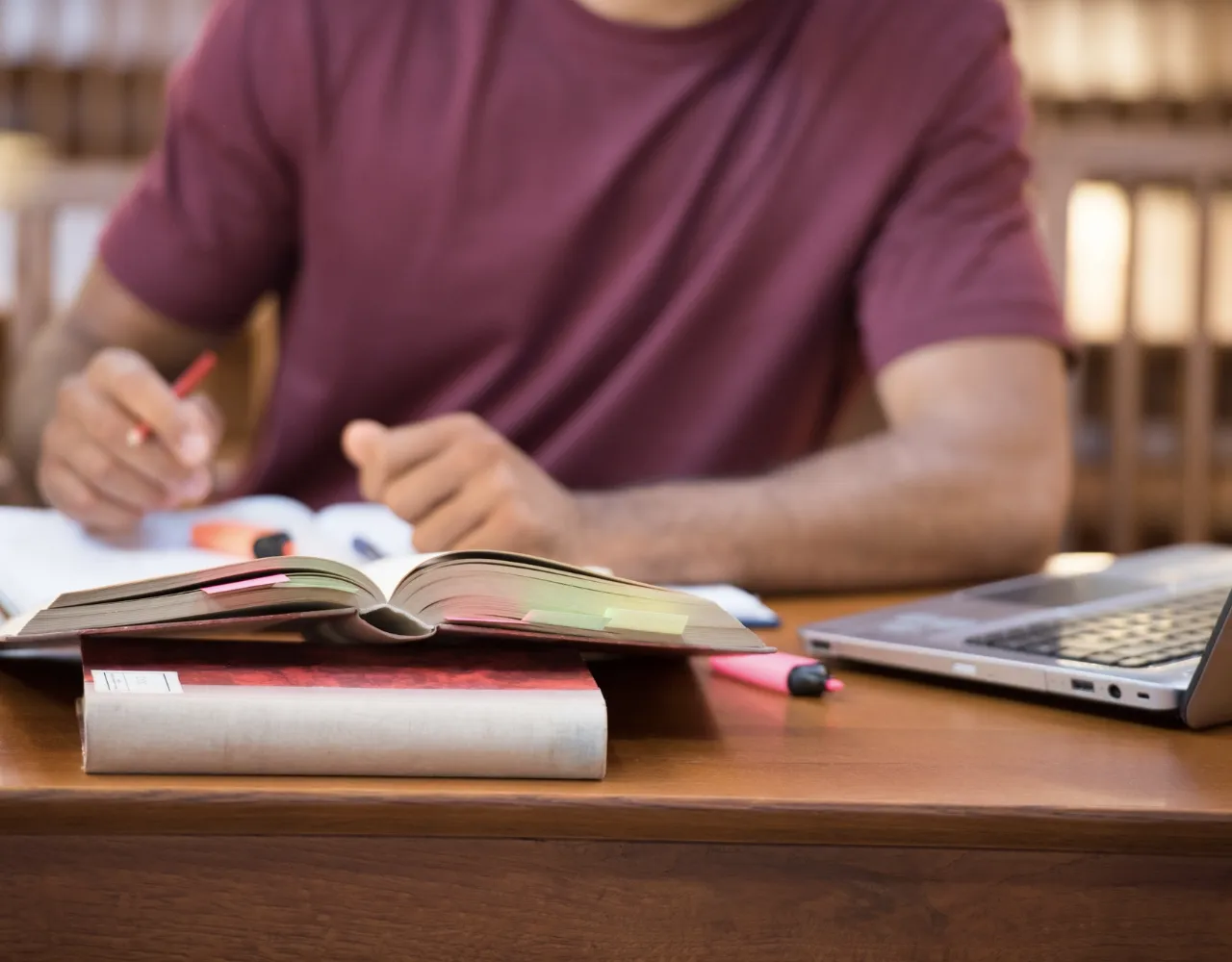 Uma pessoa de camisa marrom estuda em uma mesa com um livro, bloco de notas, marcadores de texto e um laptop.