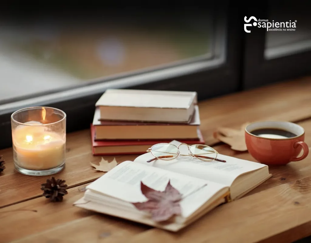 Livro aberto com uma folha sobre ele, copos, uma xícara de café, uma vela e livros empilhados sobre uma mesa de madeira perto de uma janela.