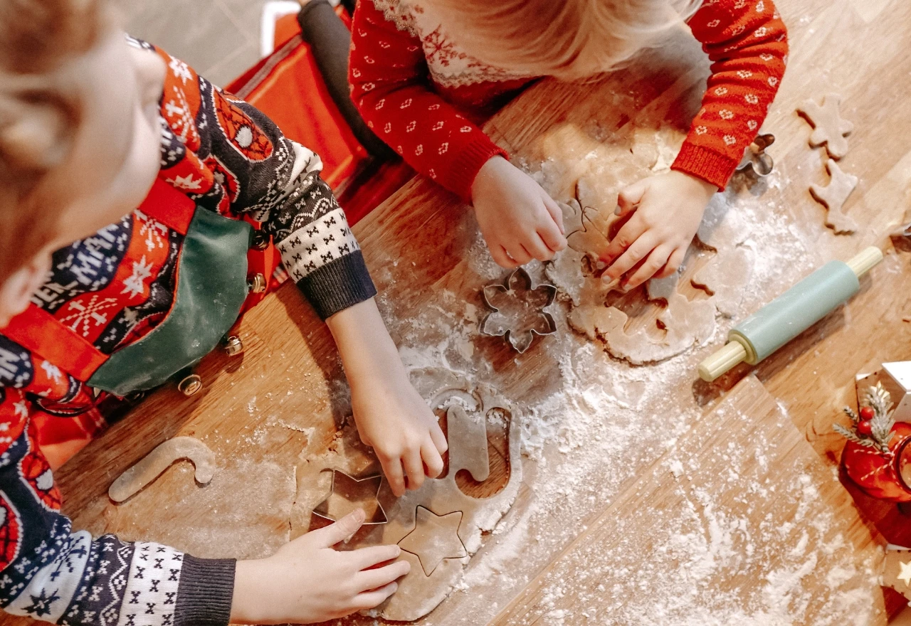 Duas crianças com suéteres festivos usam cortadores de biscoitos na massa em uma mesa de madeira, cercadas por farinha e um rolo de massa.