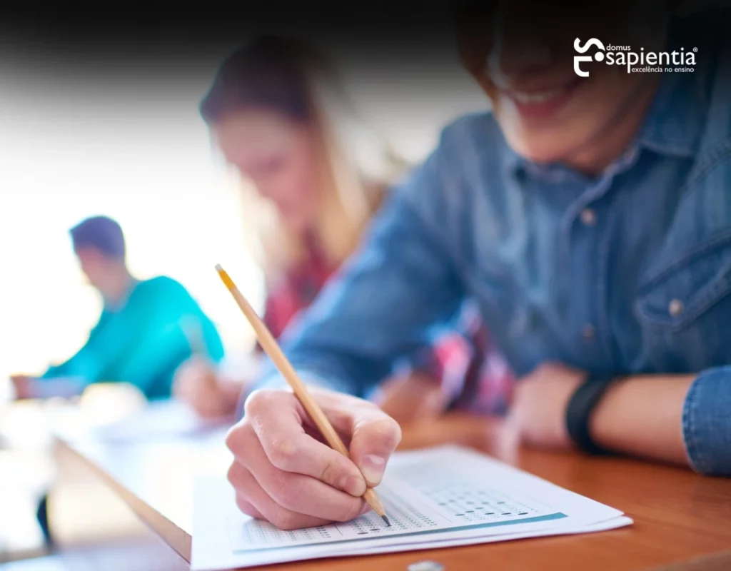 Alunos sentados em carteiras, concentrados em preencher folhas de respostas com lápis durante um teste.